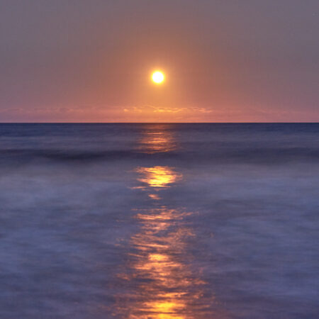Colour image of an orange full moon rising over flowing purple toned water.